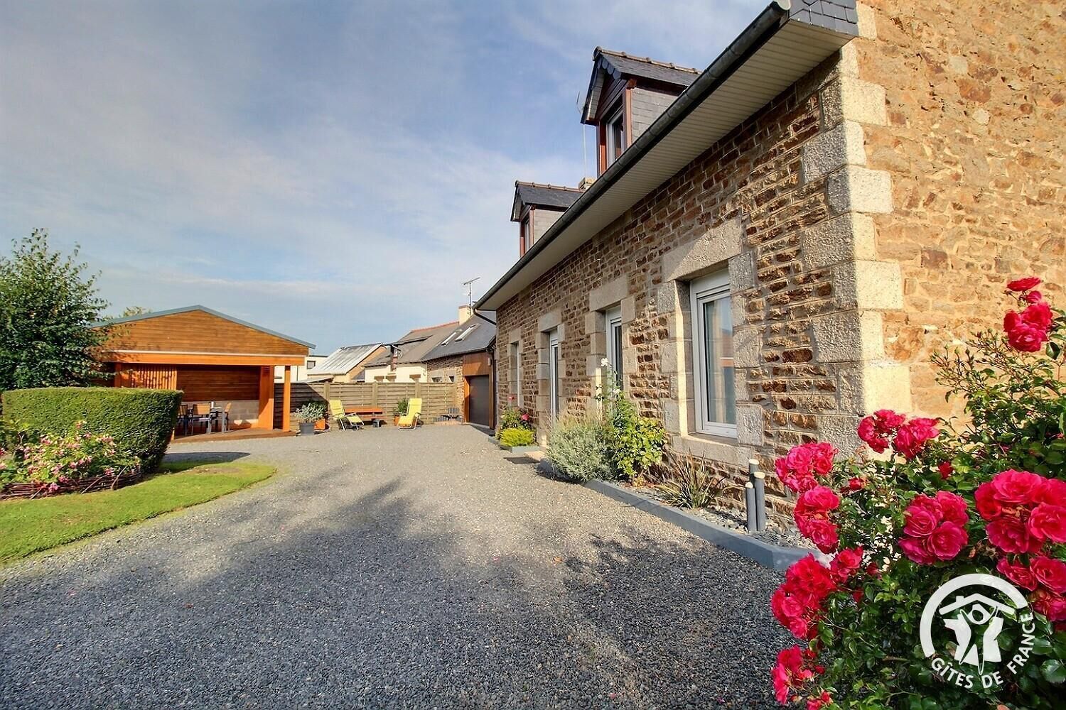 En byggnad i sten med en grusuppfart, en täckt uteplats och en trädgård med rosa blommor.