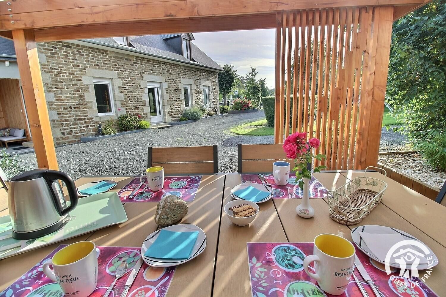 An outdoor dining area with a table set for two, a stone building in the background, and a wooden pergola.