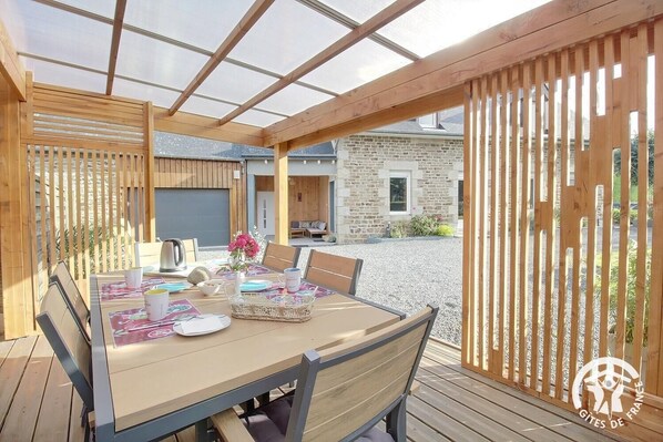 An outdoor wooden deck with a table set for dining, surrounded by wooden railings and a stone building in the background.