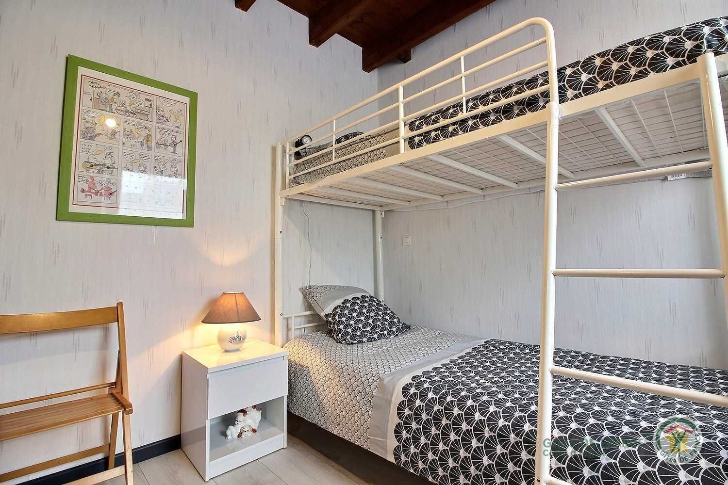 Habitación con literas, un cómic enmarcados en verde en la pared, una mesita de noche con lámpara y una silla de madera.