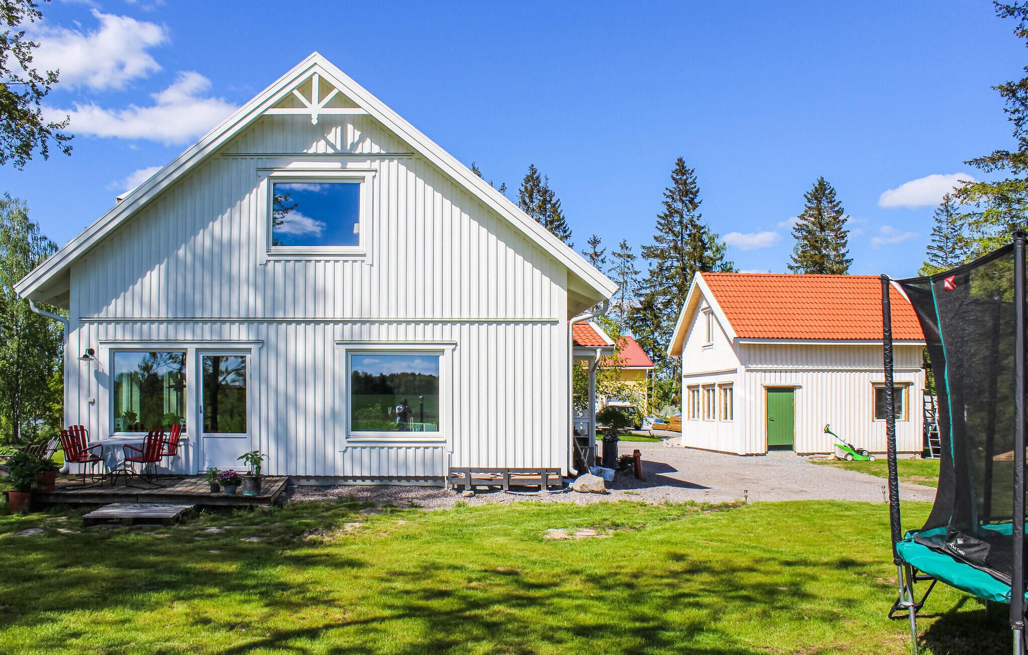 Ett vitt trähus med ett rött tak, en veranda med röda stolar och en studsmatta på gården.