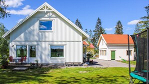 A white wooden house with a red roof, a porch with red chairs, and a trampoline in the yard.