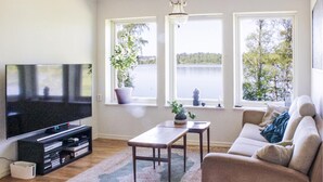 A living room with a flat-screen TV, a sofa, a coffee table, and two windows with a view of a lake.