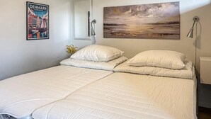 Un lit avec une literie blanche, une affiche du Danemark encadrée, un tableau représentant un paysage et un miroir.