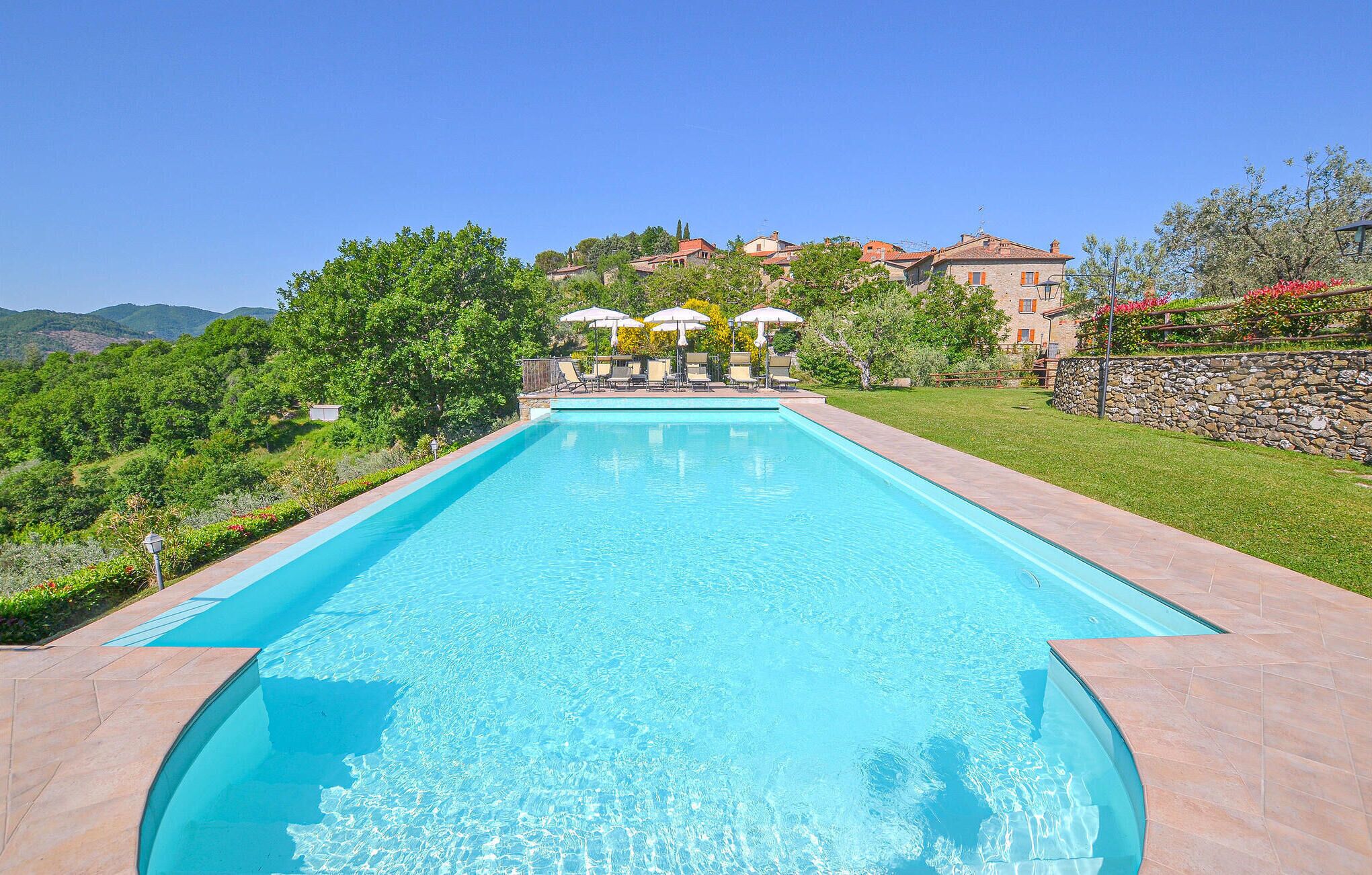 A rectangular swimming pool with clear blue water, surrounded by a stone deck. There are lounge chairs and umbrellas in the background, and a stone wall with a stone staircase leading to a building.