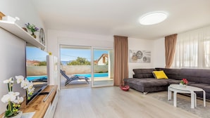 A modern living room with a grey sofa, a small white table, and a TV on a wooden stand. There is a sliding glass door leading to a balcony with a pool. - Cozy home in Linardici (Linardici)