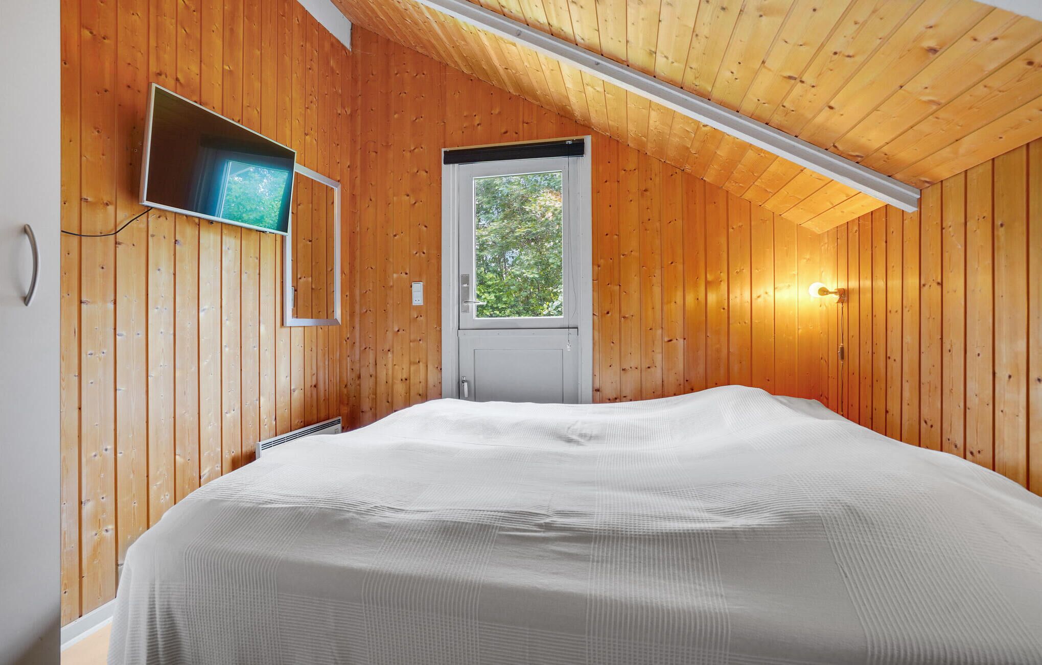 Un dormitorio con paredes de madera, una cama con cubrecama blanco, un televisor montado en la pared y una ventana con vistas a árboles.
