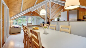 A spacious kitchen with a high ceiling and wooden beams, a dining area with a white table and wooden chairs, and a view of the outdoors through large windows. - Beautiful home in Sydals with sauna (Sydals)