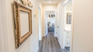 Un couloir avec un plancher en bois, un grand cadre orné sur le mur de gauche et un lit avec un panneau indiquant « LU » en arrière-plan.
