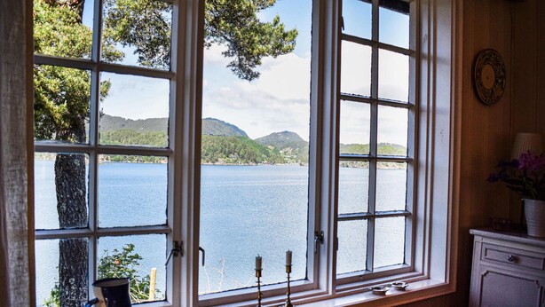 Ein Zimmer mit großem Fenster mit Blick auf einen See, einem Holzschrank und einer Vase auf der Fensterbank.