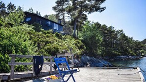 Eine Holzstege mit einer Bank und einem Stuhl, ein Metallkübel mit Blumen, und ein Gebäude mit blauen Fenstern auf einer Anhöhe.