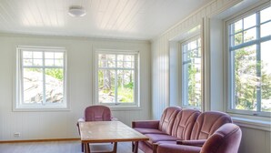 Een kamer met een houten tafel en twee comfortabele stoelen, grote ramen met uitzicht op bomen en een gebouw, en witte wandpanelen.