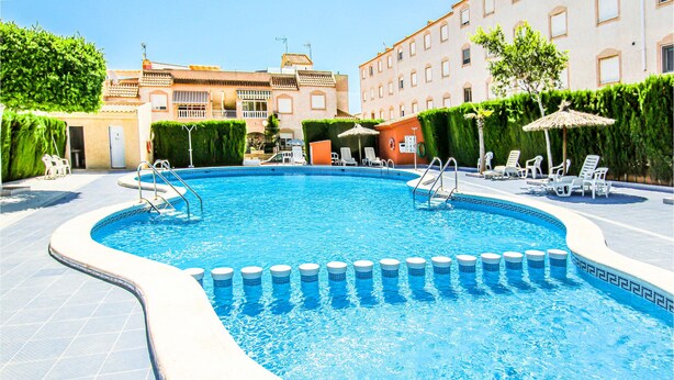 A swimming pool with a tiled deck, surrounded by lounge chairs and umbrellas, with buildings in the background.