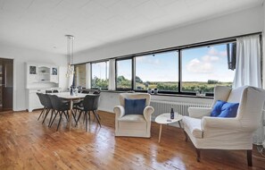A spacious living area with a dining table, two armchairs, and a sofa. A large window offers a view of greenery outside.