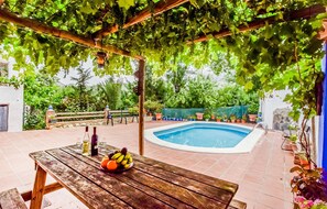 Une table en bois sur laquelle se trouvent un panier de fruits et des bouteilles de vin, une piscine et une pergola couverte de vignes.