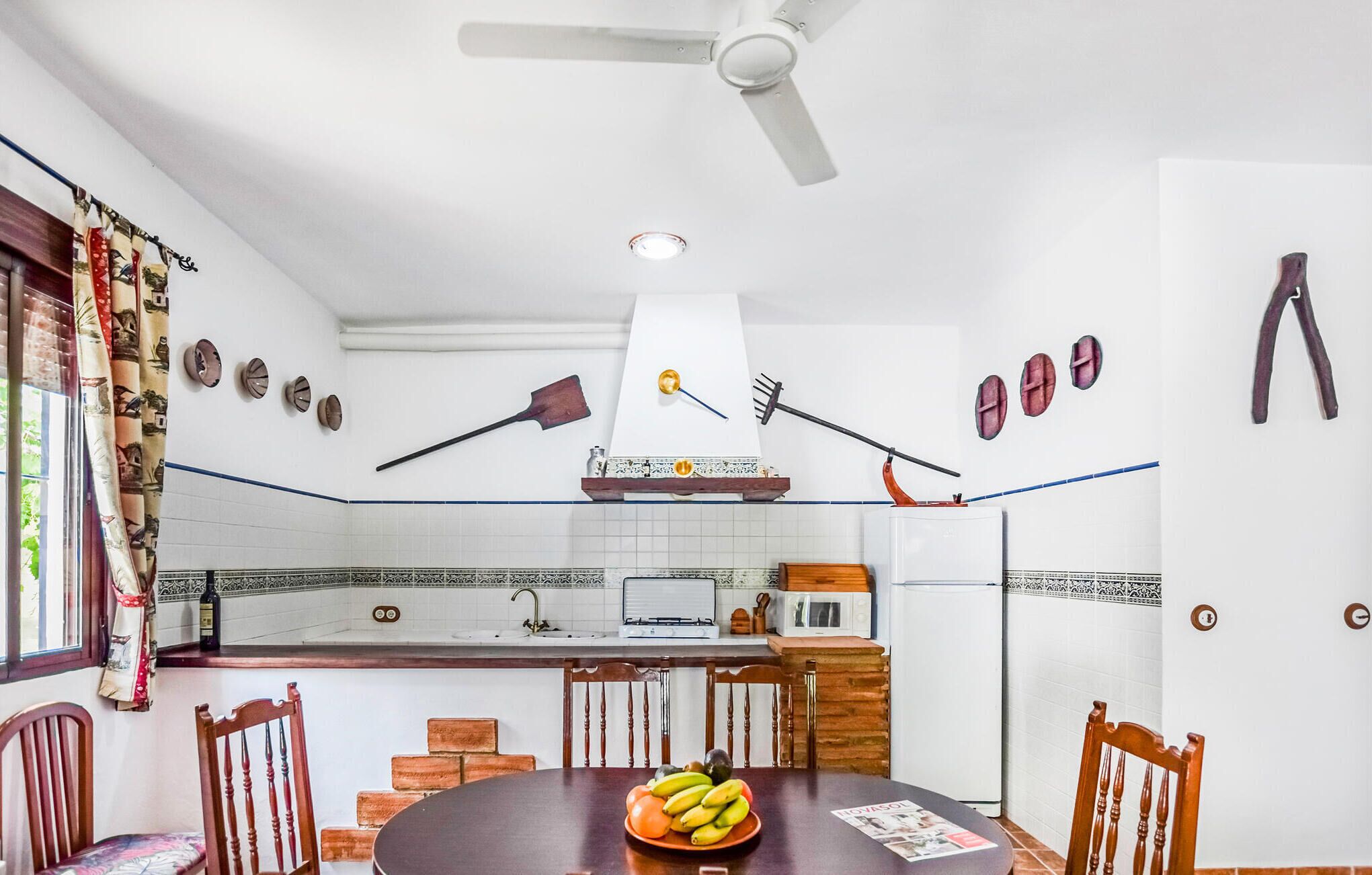 Cuina amb sostre blanc, ventilador de sostre, un rentaplat, fogó i microones. Hi ha cadires de fusta i una taula amb un plat de fruita.