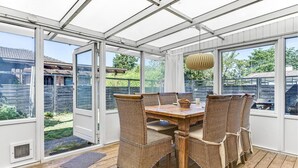 Une véranda avec une table en bois et des chaises en osier, de grandes fenêtres et un luminaire suspendu.