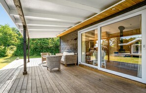 En overdækket terrasse med fletmøbler, et træterrasse og en glasdør, der fører til et indre rum.