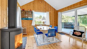 A cozy room with a wood-burning stove, a dining table with blue chairs, and a large window offering a view of the outdoors.