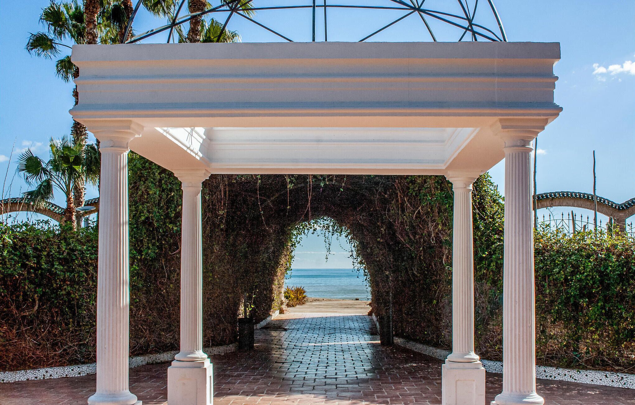 Eine weiße Pergola mit Säulen, die zum Strand führt.