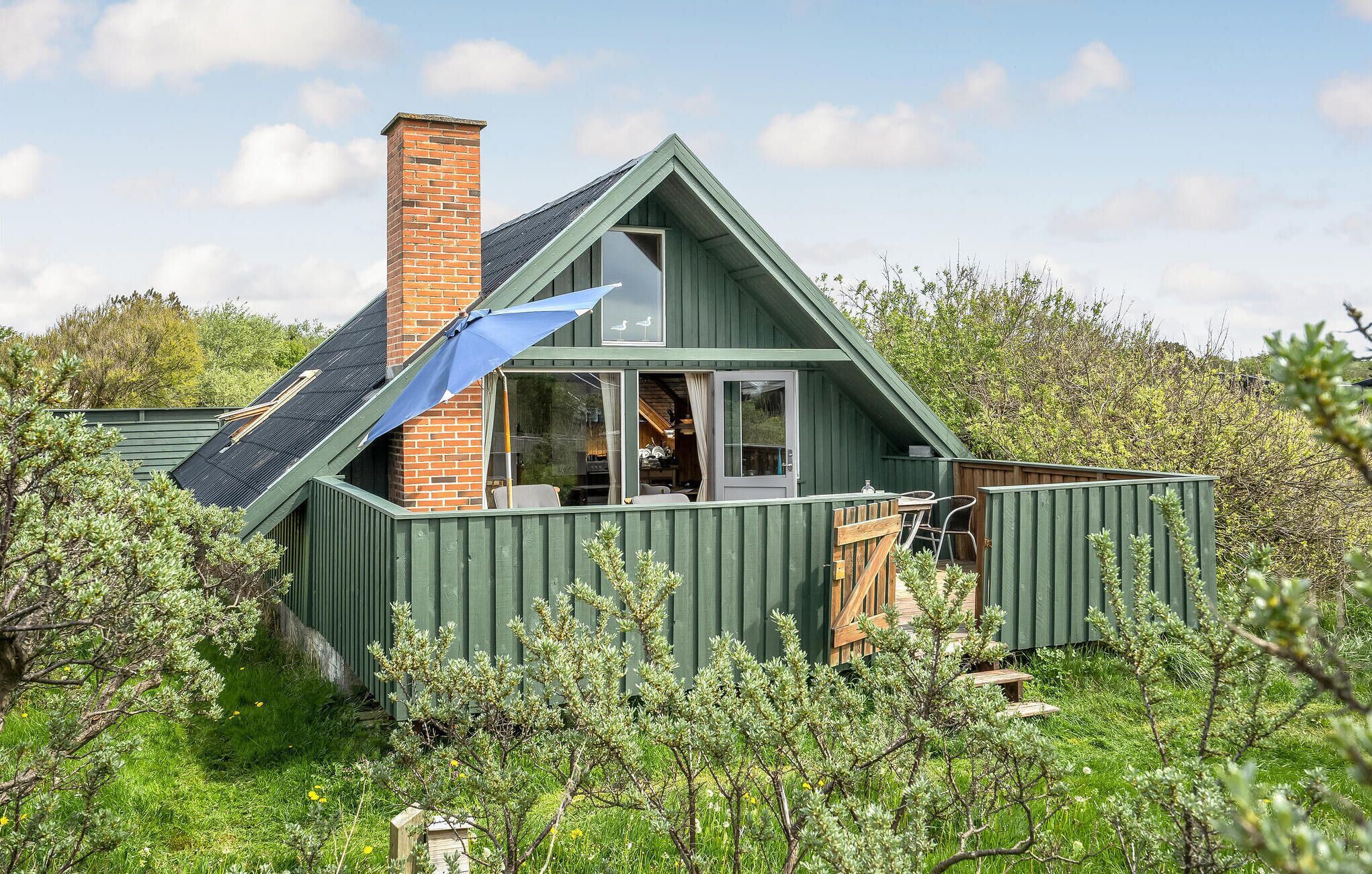 A small, green-roofed cabin with a brick chimney, surrounded by lush greenery.