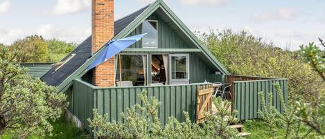 A small, green-roofed cabin with a brick chimney, surrounded by lush greenery.