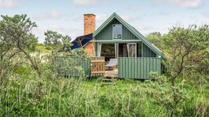 A small, green-roofed cabin with a brick chimney, a deck with a lounge chair, and a large window. - Amazing home in Sjællands Odde (Sjællands Odde)