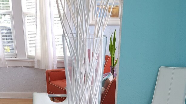 A white wicker table with a glass top, surrounded by white chairs, in a room with a blue wall and a window with white curtains.