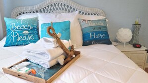 A neatly made bed with a tray of towels and toiletries, two pillows with 'Beach is my Happy Place' text, and a bedside table with a clock and a vase.