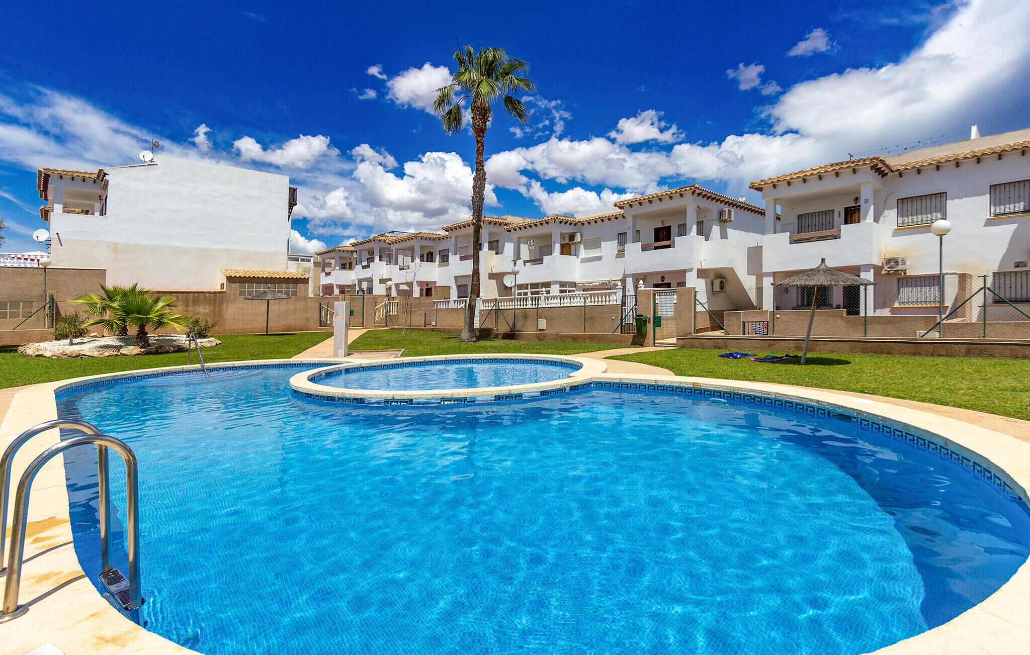 A swimming pool surrounded by white buildings with terracotta roofs, a palm tree, and a clear blue sky.