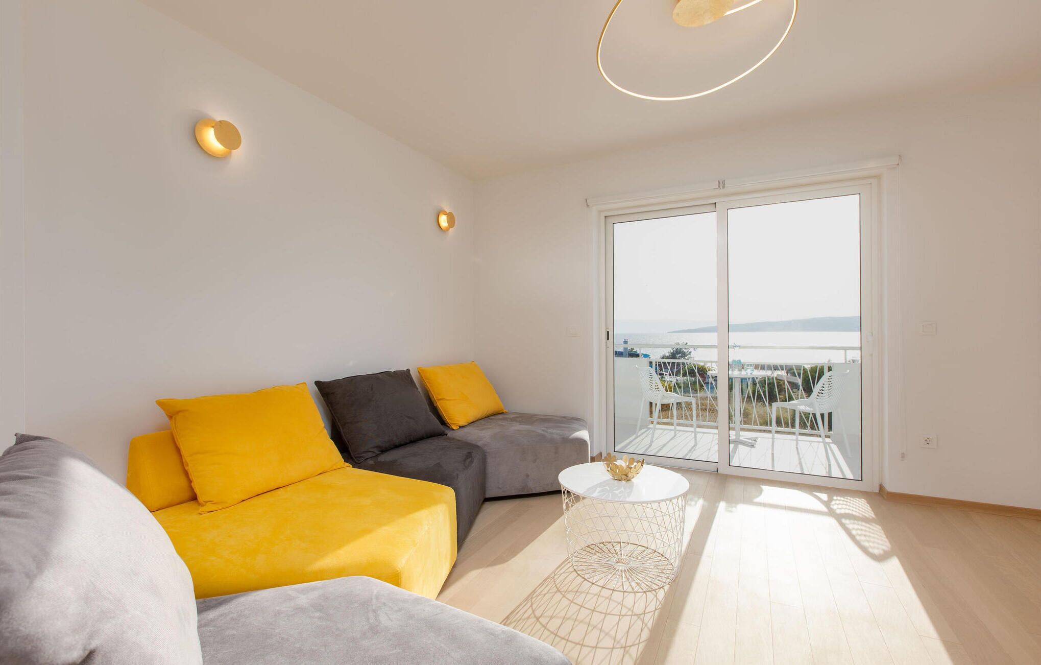 Una sala de estar moderna con un sofá gris y cojines amarillos, una mesa de centro blanca y un amplio ventanal corredero que da a un balcón con vistas.