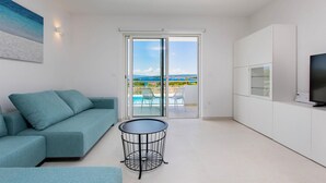 A modern living room with a teal sofa, a small round table, and a glass door leading to a balcony with a pool and a scenic view. - Awesome home in Punat (Punat)
