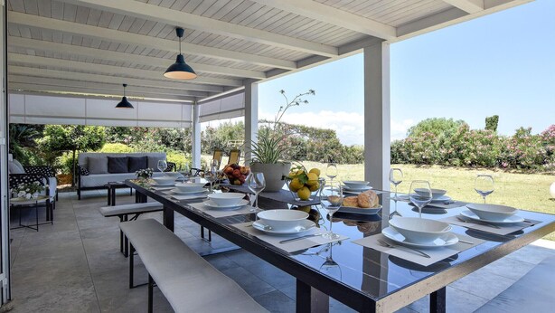 Un espace de restauration en plein air avec une longue table dressée pour un repas, des bancs et une vue sur le jardin.