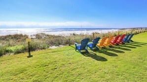 A grassy area with a row of colorful Adirondack chairs facing a beach.