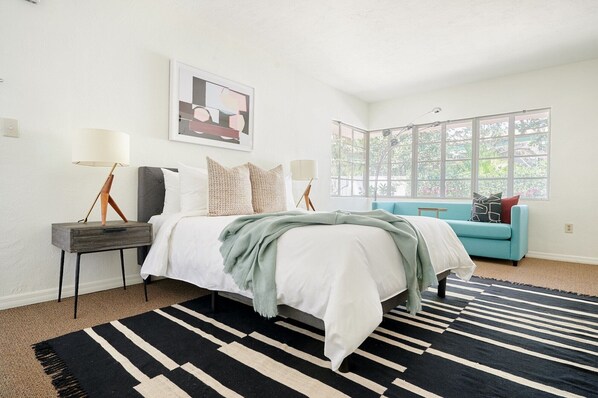 A bedroom with a bed, bedside tables, a blue sofa, a large window, and a black and white striped rug. - Courtyard at Lake Lucerne (Orlando)