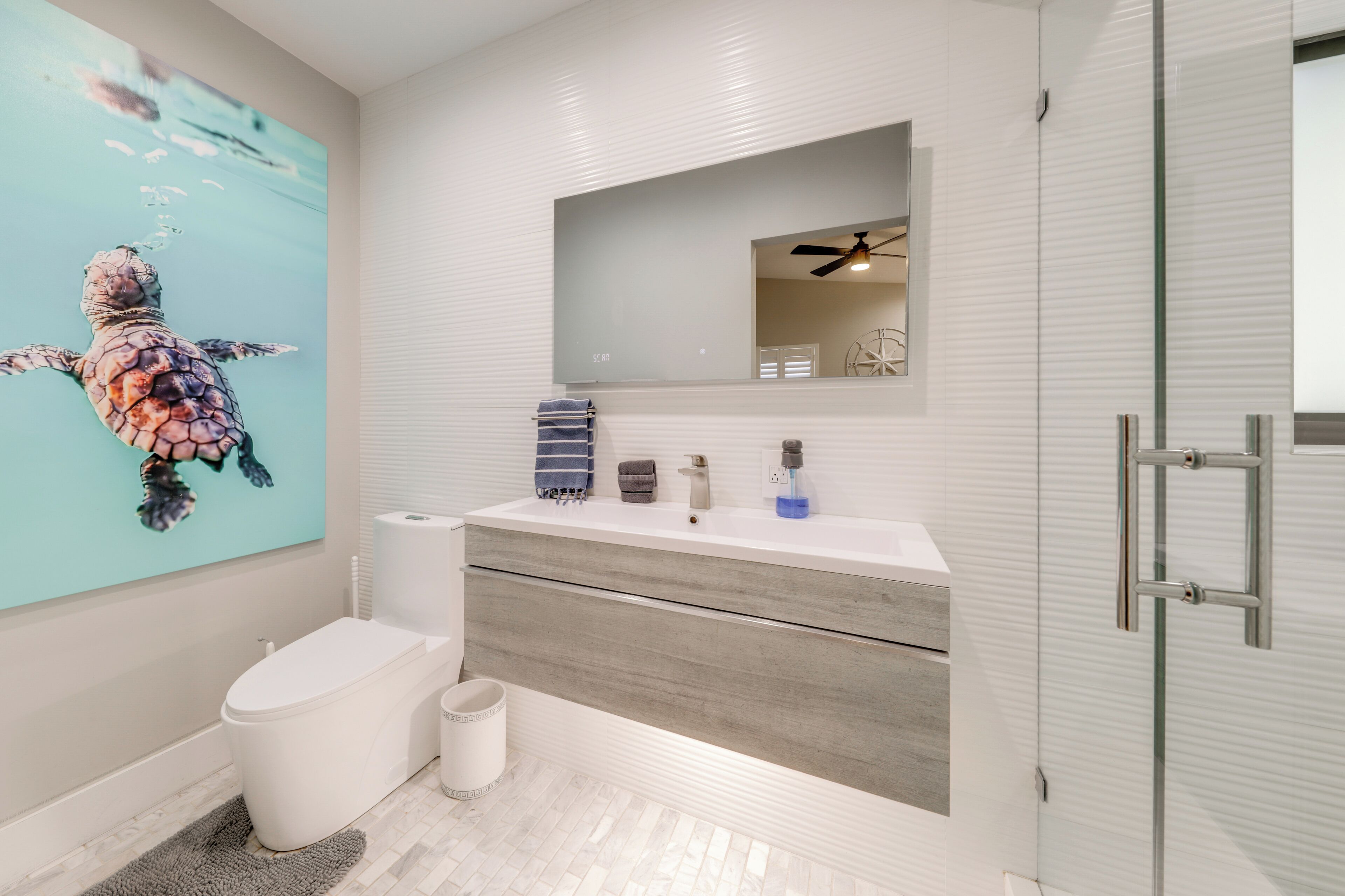 A modern bathroom with a white vanity, a large mirror, a ceiling fan, and a wall-mounted toilet.