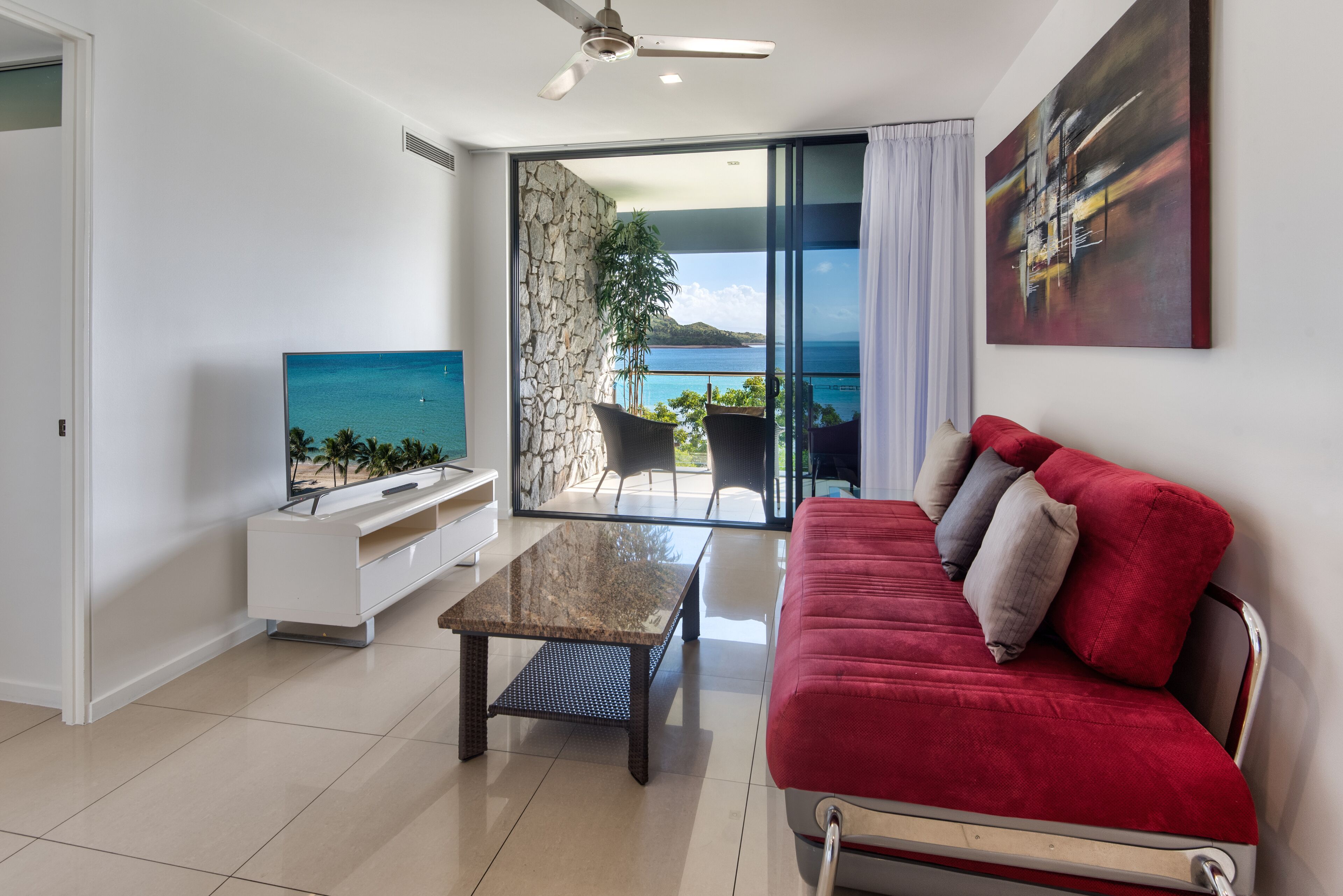 A modern living room with a TV, a red sofa, a coffee table, and a view of the sea through a glass door.