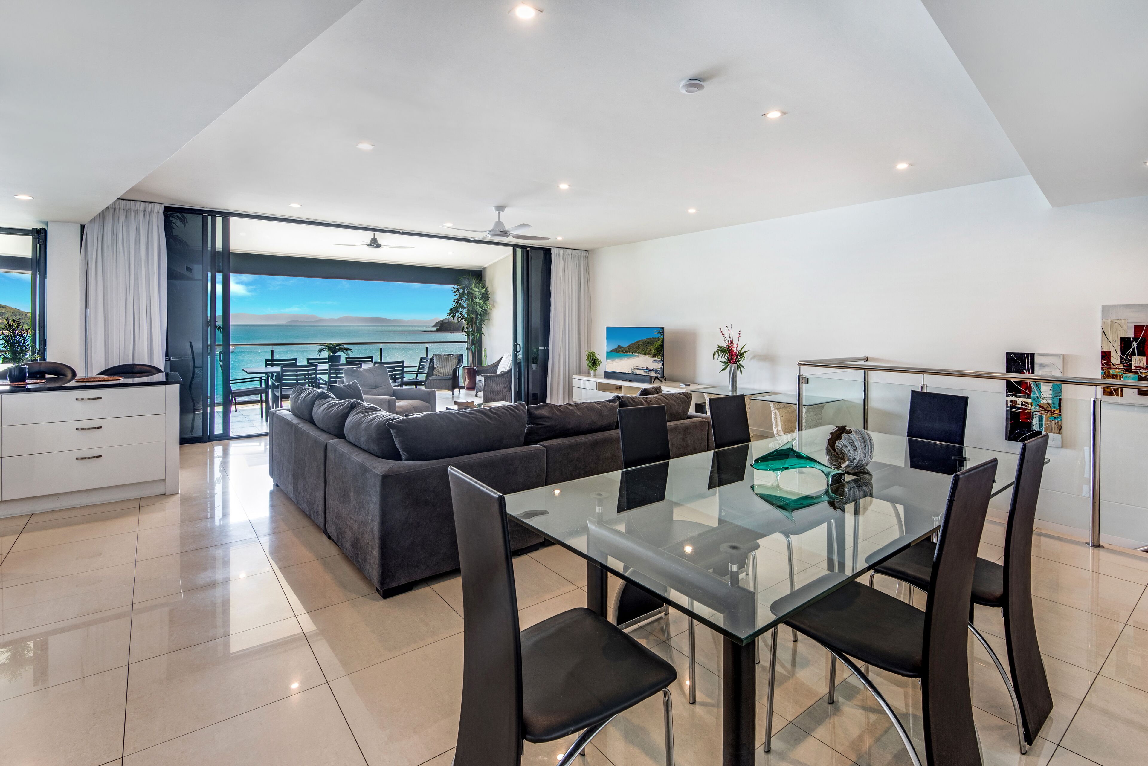 Uma sala de estar moderna com uma mesa de jantar de vidro, sofás escuros e vista para o mar.