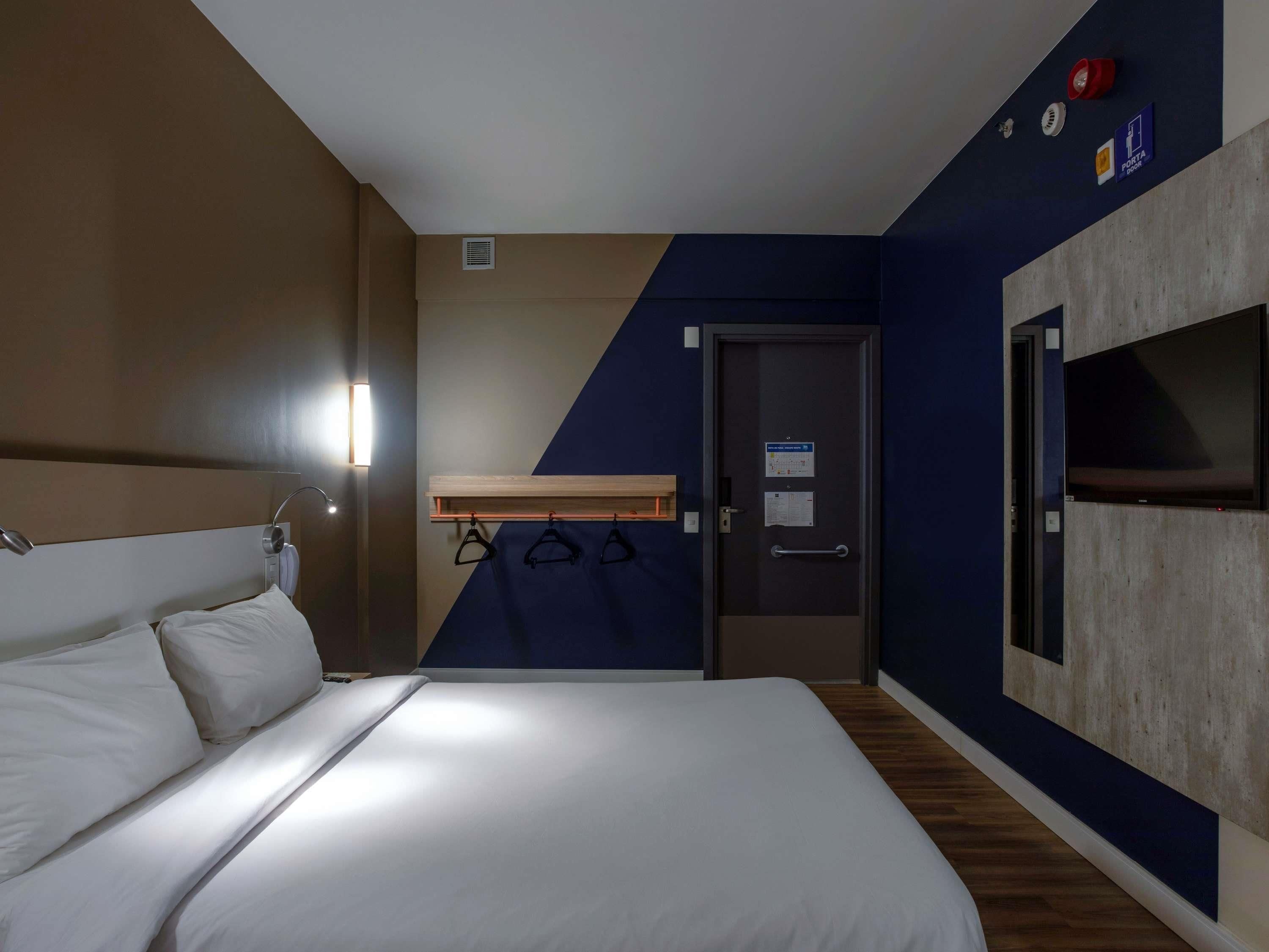 A hotel room with a bed, a TV mounted on the wall, a coat rack, and a door with a sign above it.