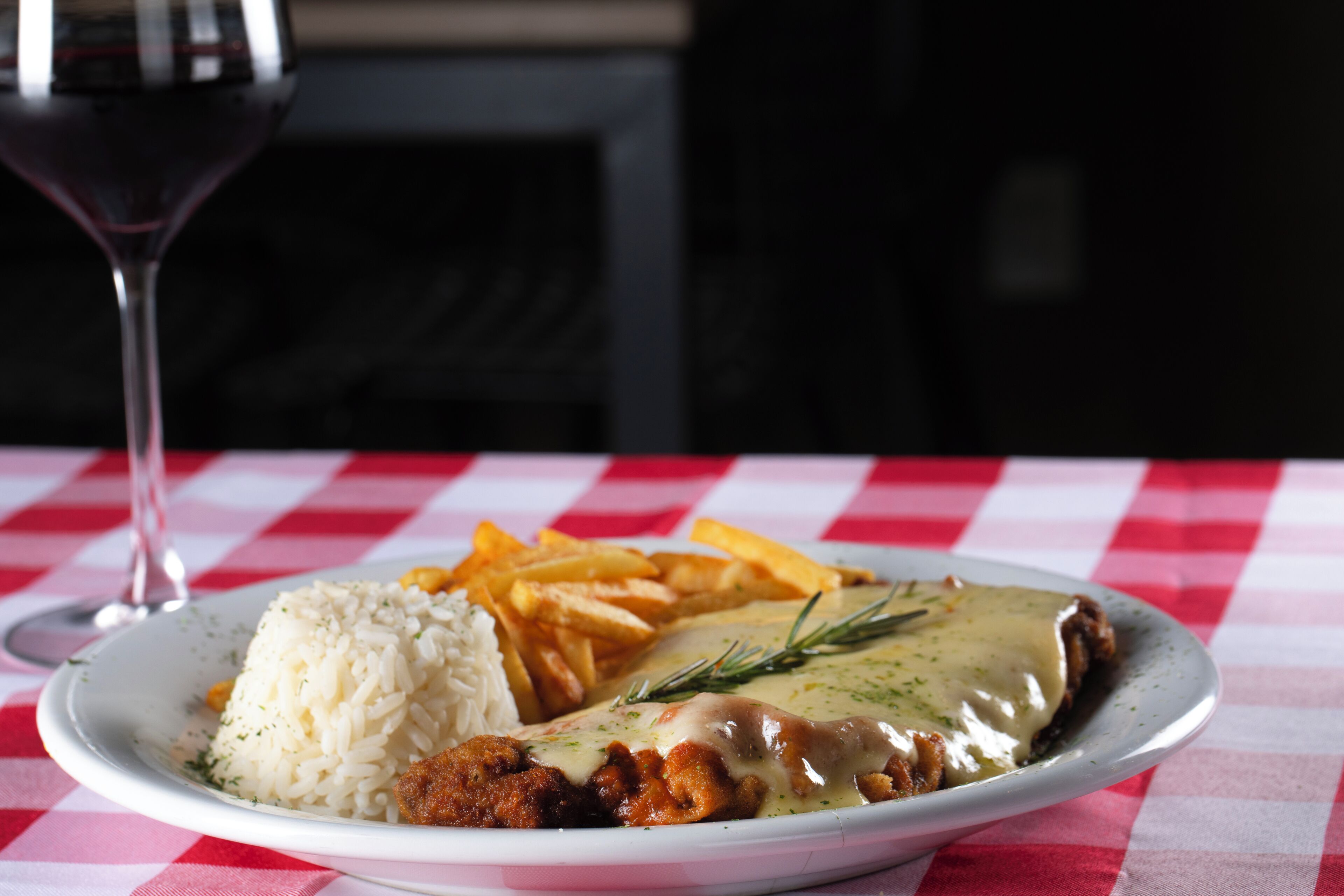 A plate of food with rice, a meat dish, and fries, accompanied by a glass of red wine on a checkered tablecloth.