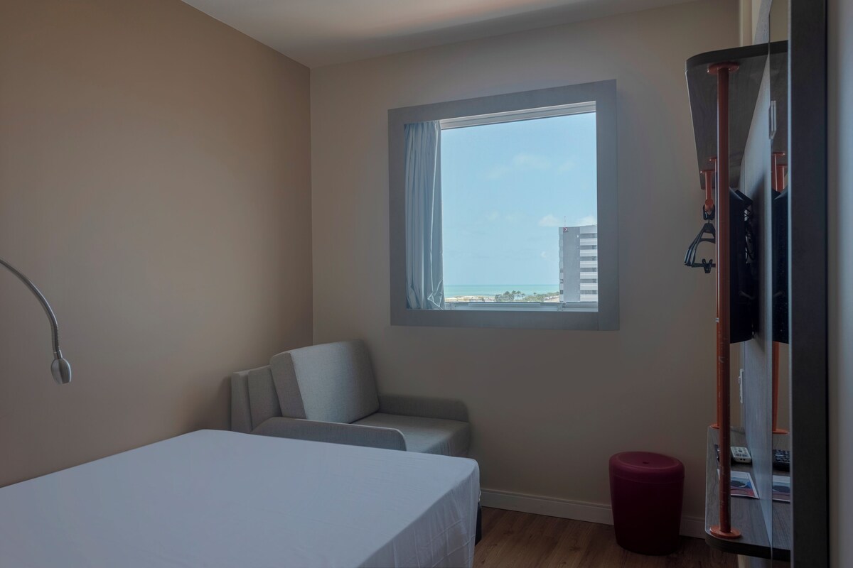 Une chambre d’hôtel avec un lit, un petit canapé, une fenêtre donnant sur un immeuble et une poubelle rouge.