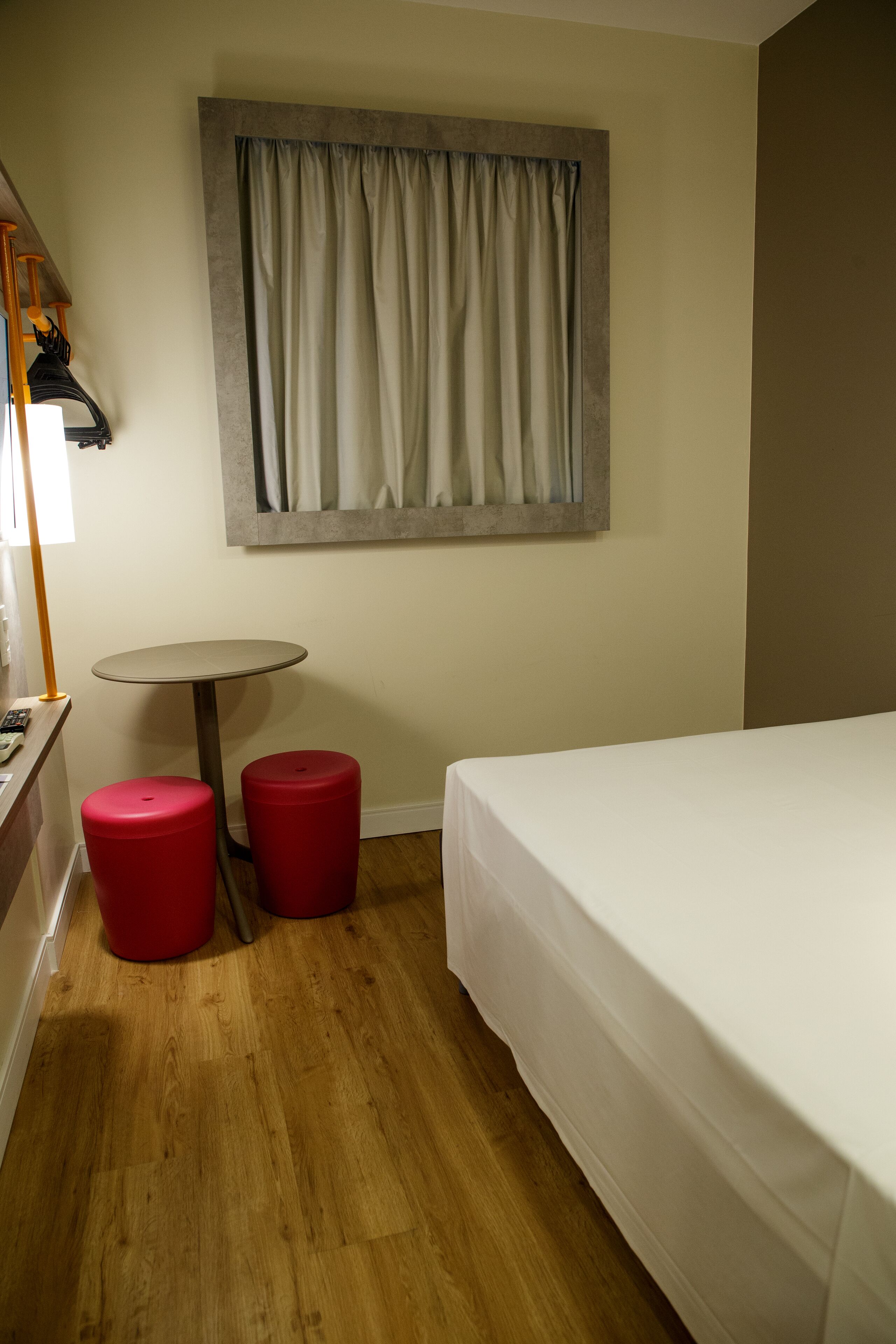 A hotel room with a bed, a small round table, two red stools, a lamp, and a window with curtains.