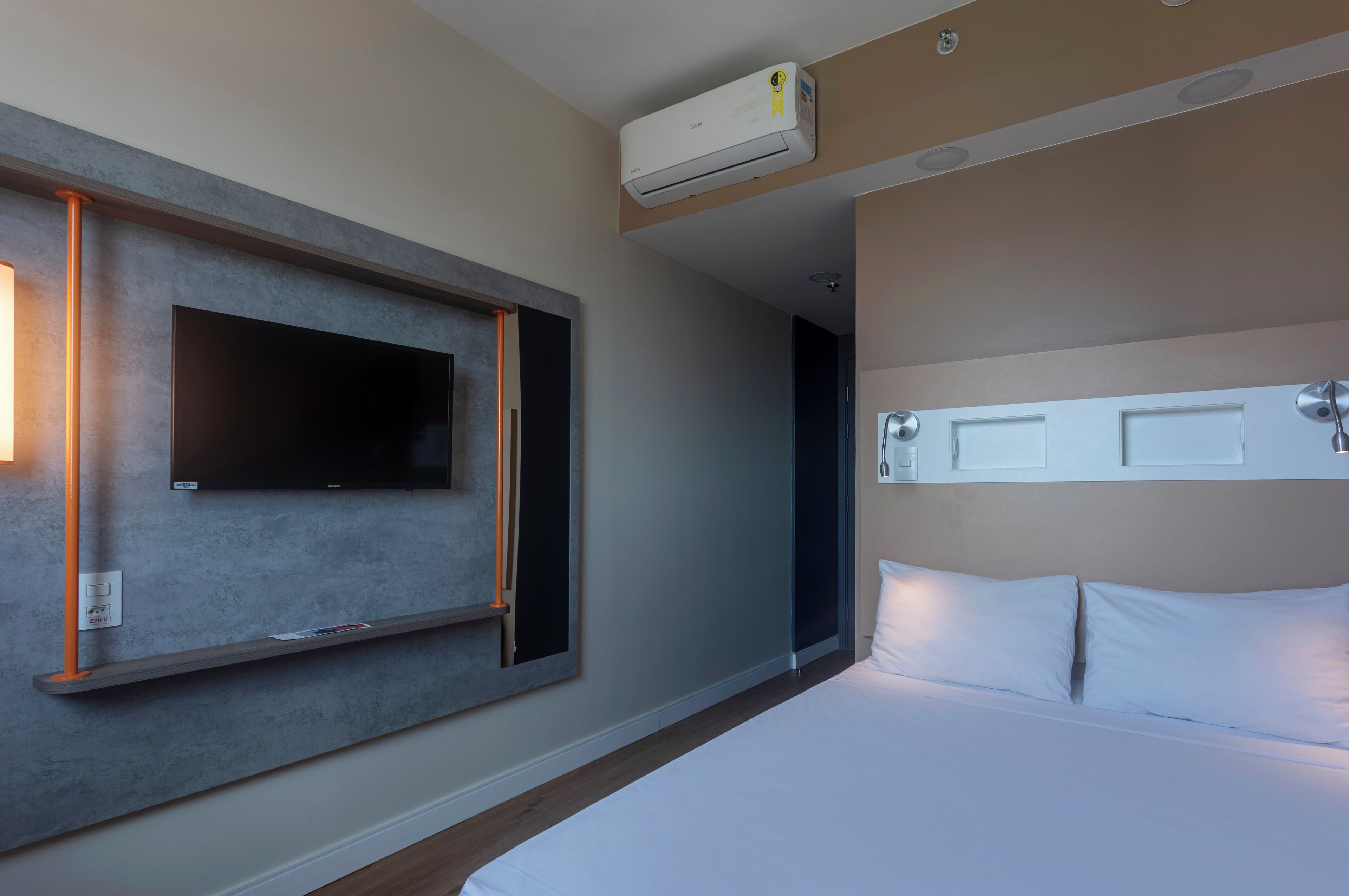 Une chambre d’hôtel dotée d’un téléviseur à écran plat fixé au mur, d’un lit avec du linge de lit blanc, d’un climatiseur fixé au plafond et d’une porte avec une poignée.