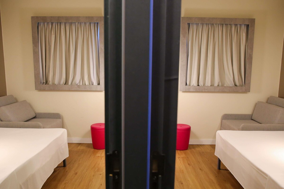 A hotel room with two beds, a red stool, and curtains on the windows.