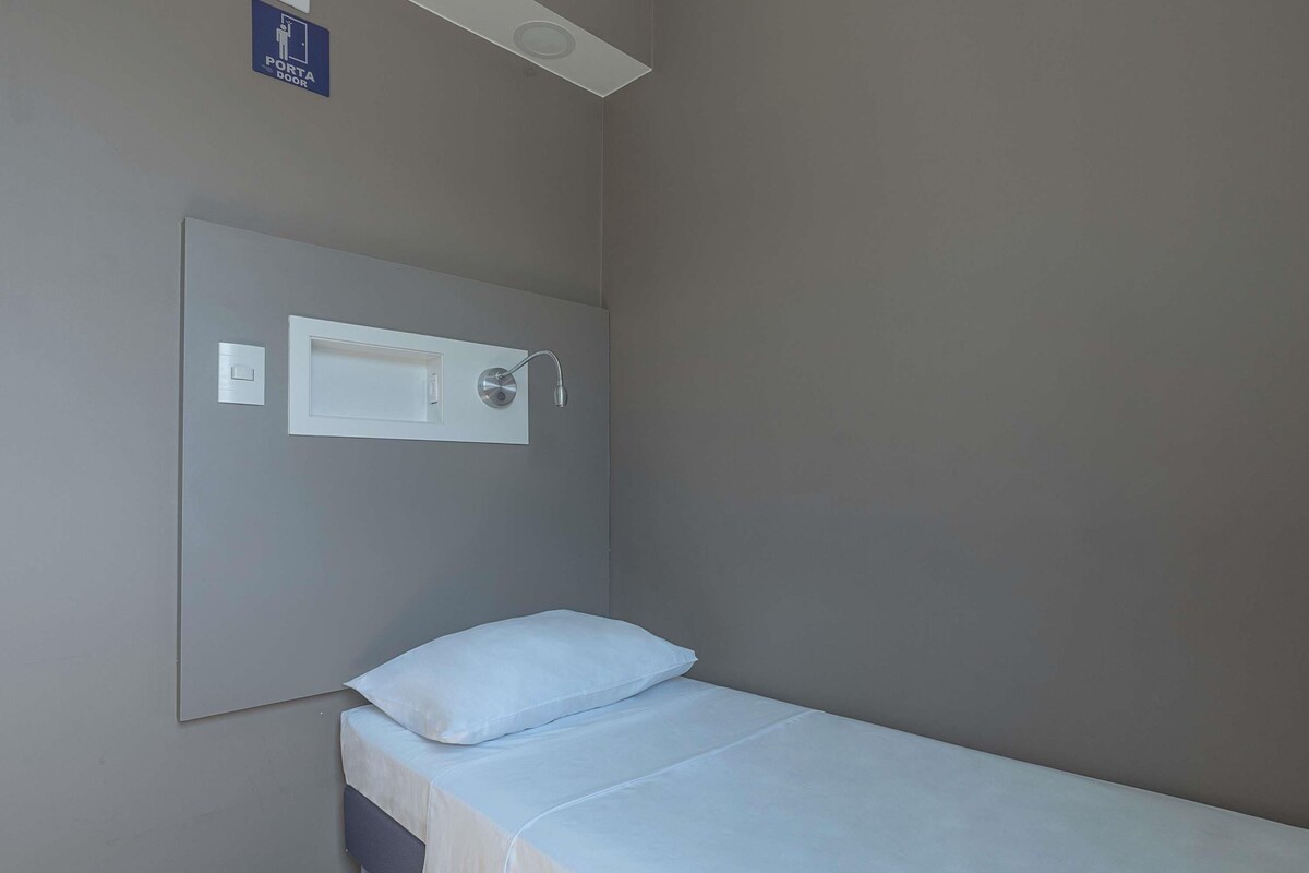 A small, single-bed room with a headboard, a wall-mounted light, and a door with a sign above it.