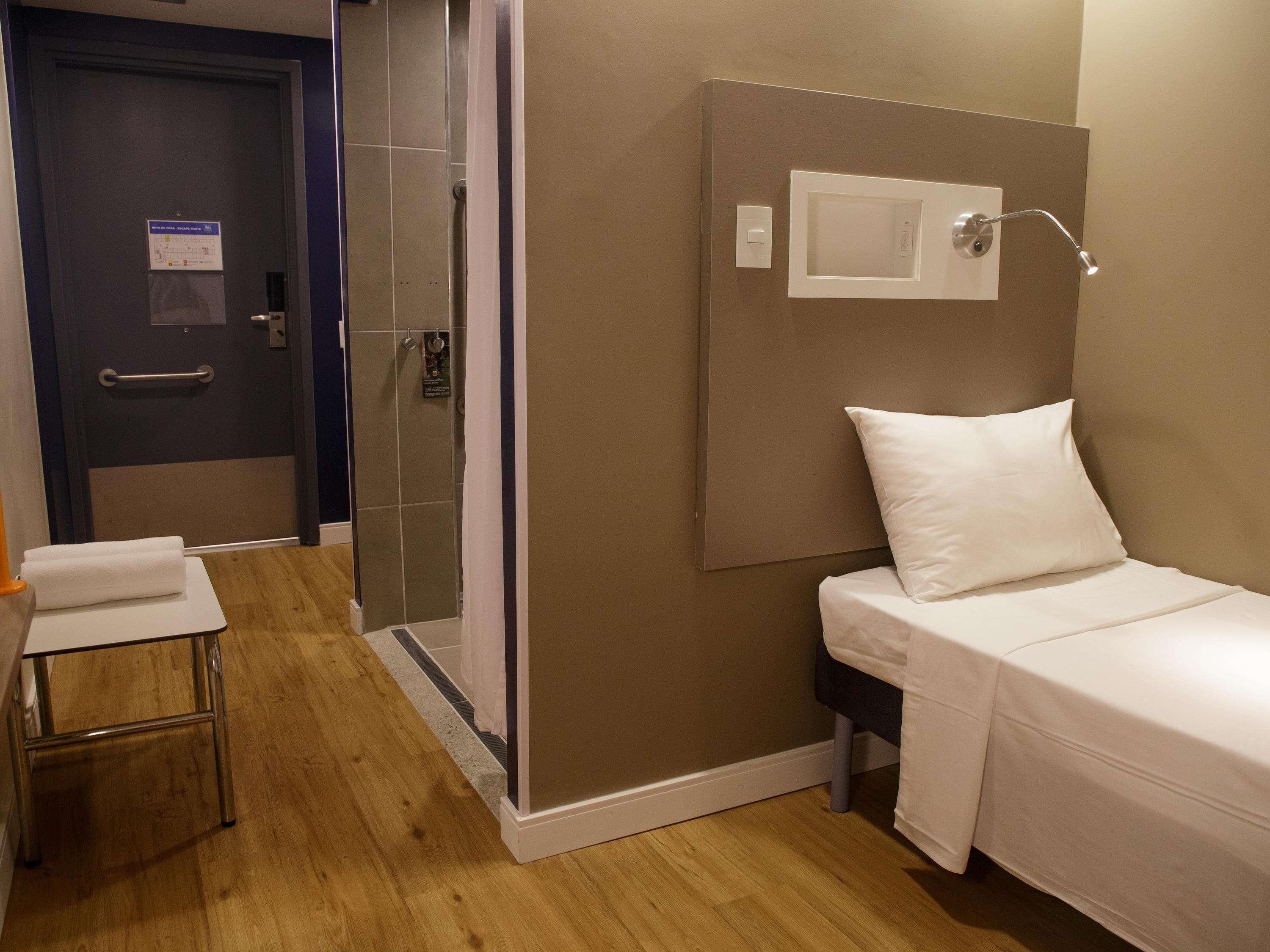 A hotel room with a bed, a bedside table, a shower area, and a door with a handle.