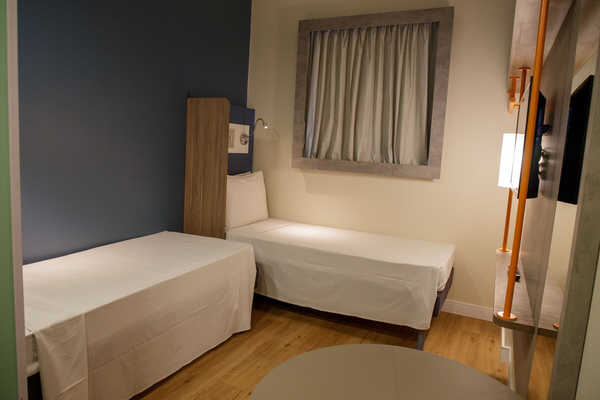 Une petite chambre à coucher, bien rangée, avec deux lits simples, une armoire en bois, une fenêtre avec des rideaux et une table de chevet avec une lampe.