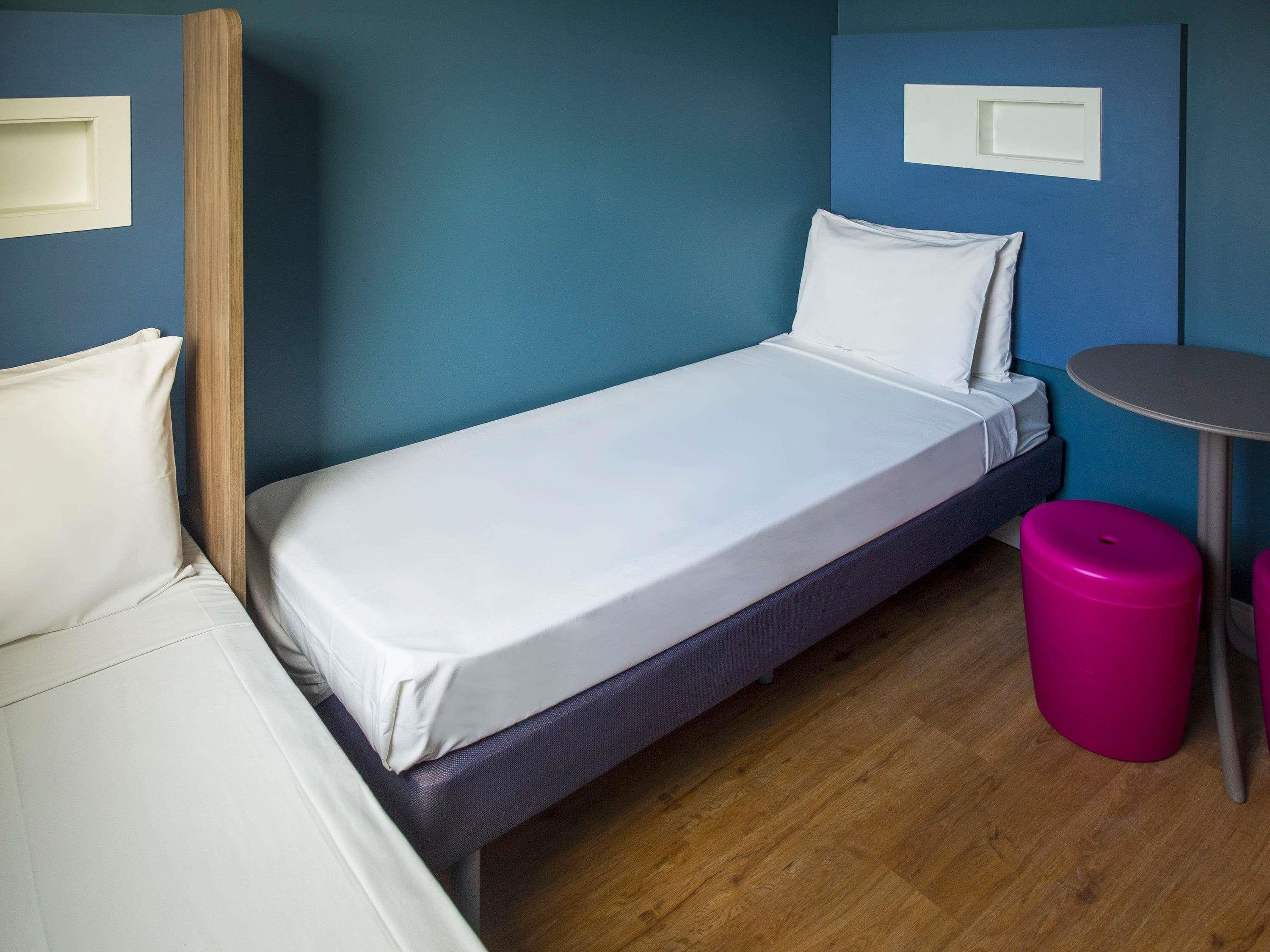 Een enkel bed met wit beddengoed, een klein rond tafeltje, een roze kruk en een houten vloer.