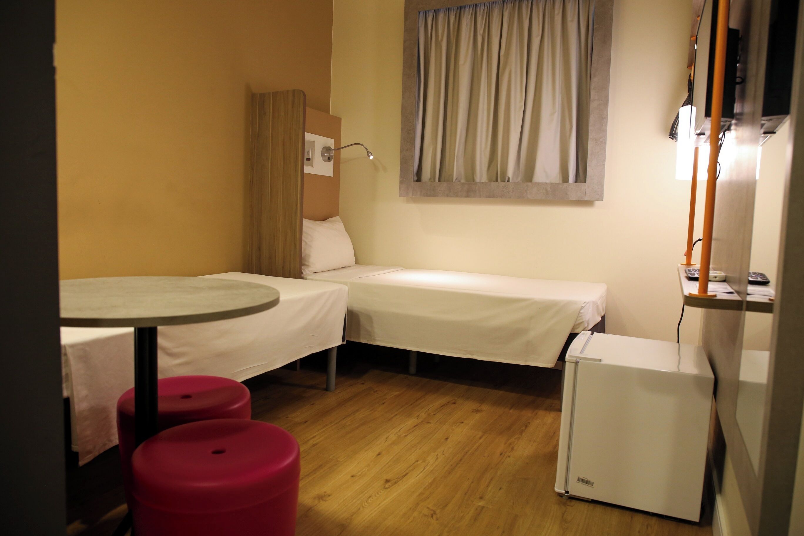 Una habitación de hotel pequeña con dos camas individuales, una mesita, un refrigerador y una ventana con cortinas.