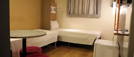 Una habitación de hotel pequeña con dos camas individuales, una mesita, un refrigerador y una ventana con cortinas.
